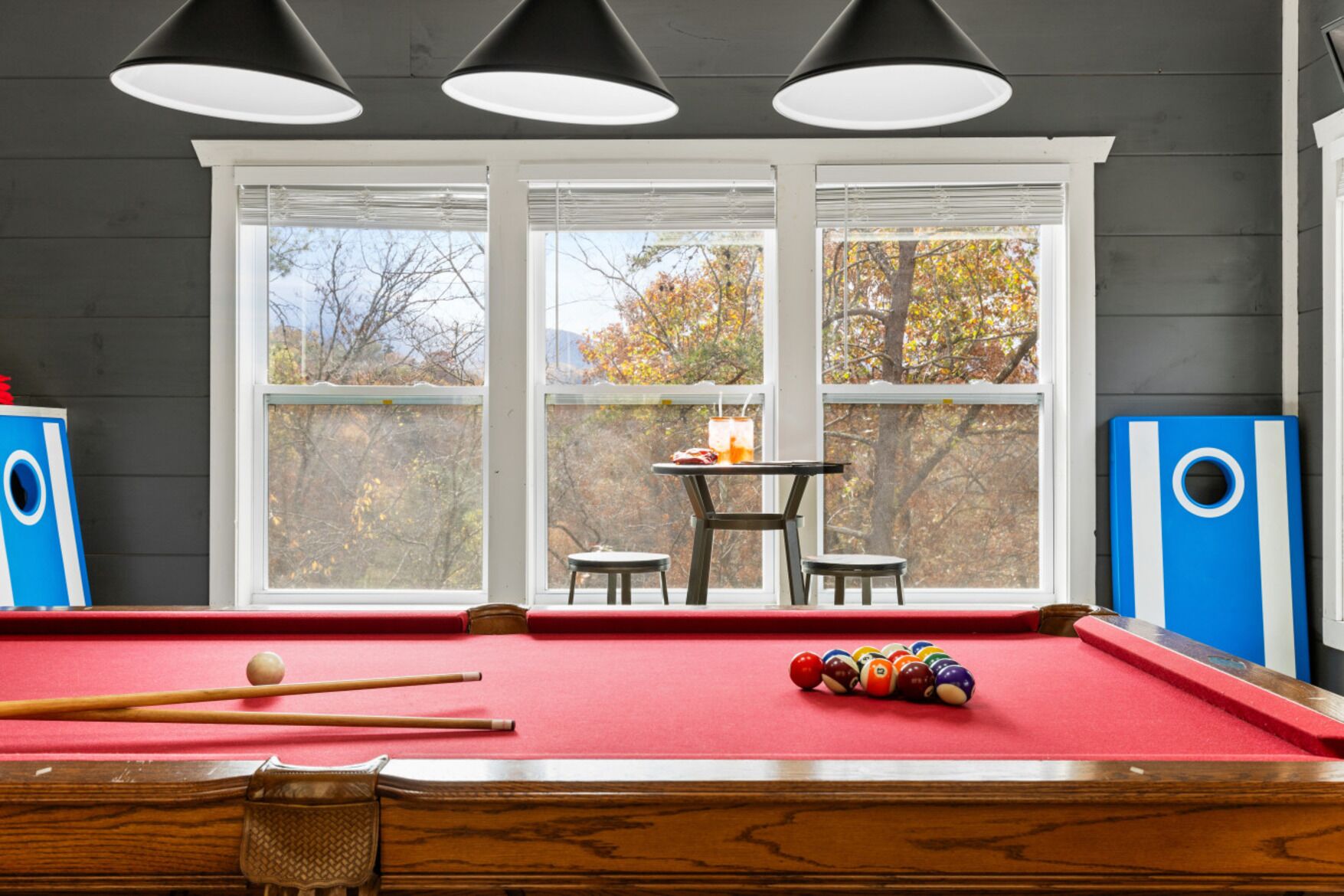 Ein Billardtisch mit roter Spielbahn, zwei Billardstöcke und ein Satz farbiger Kugeln. Darüber hängen drei schwarze Pendelleuchten. Im Hintergrund ist ein Fenster mit weißen Rahmen zu sehen, durch das Herbstlaub sichtbar ist.