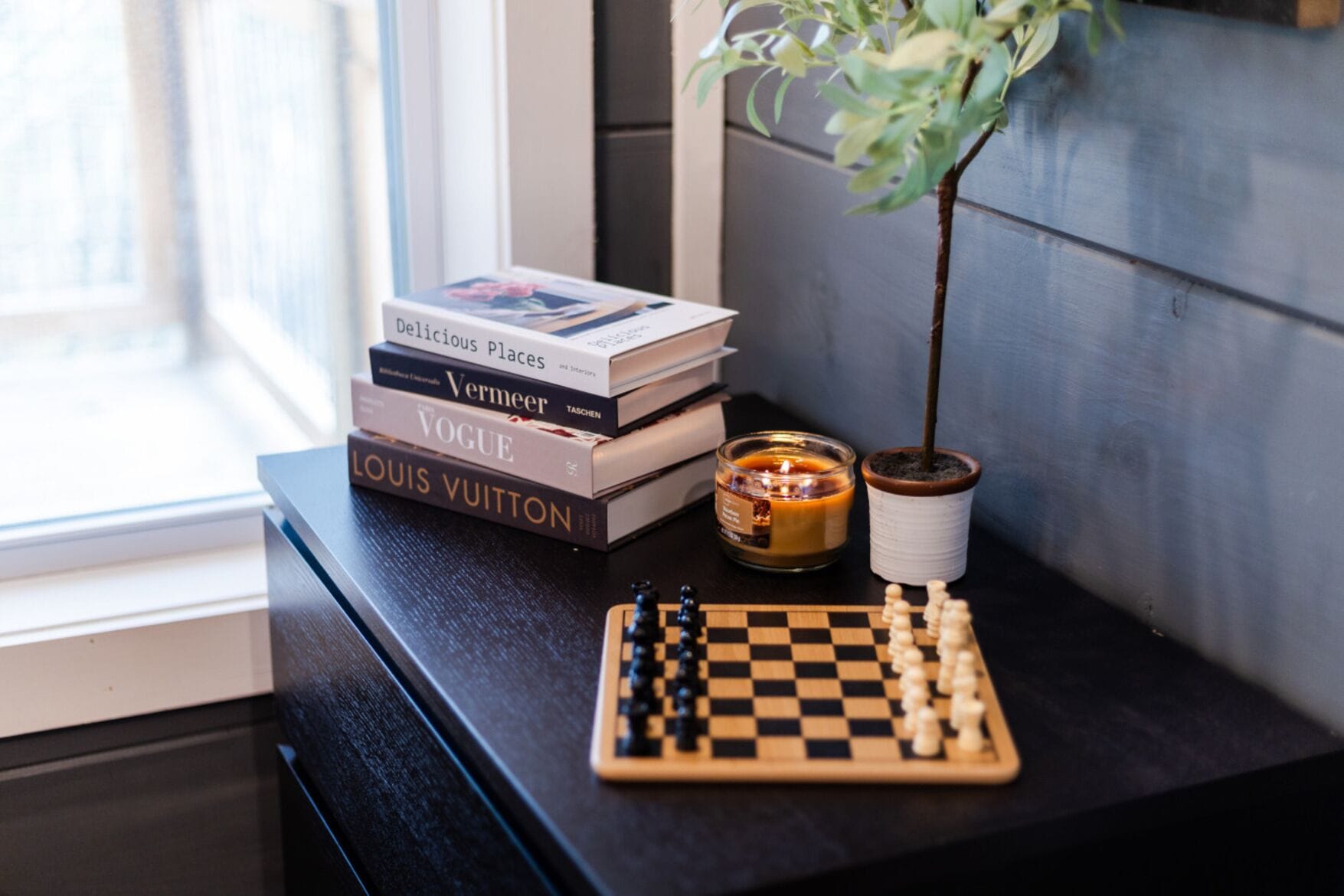 A stack of books, a candle, and a chessboard on a dark surface.