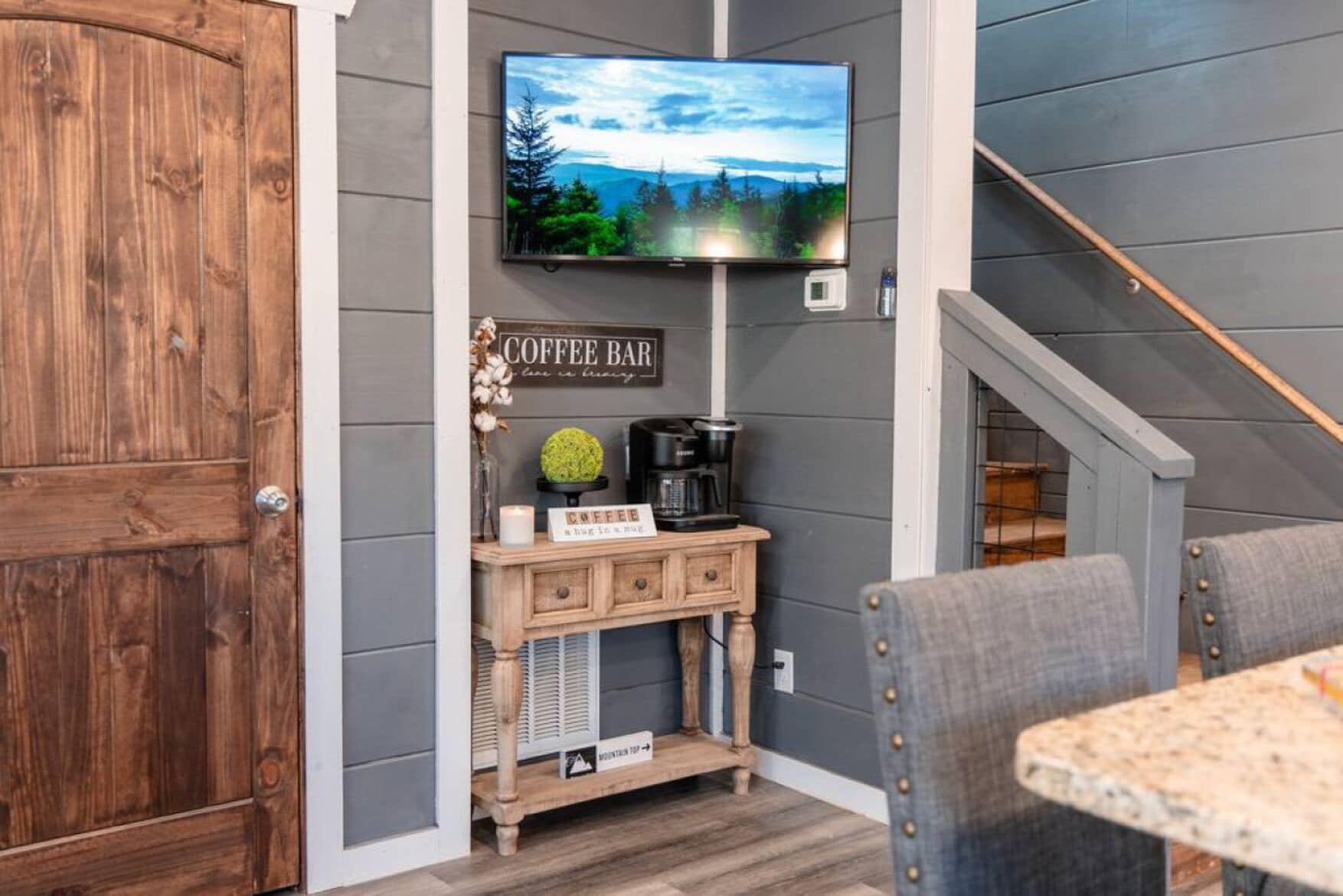 Una habitación acogedora con puerta de madera, una barra de café con un cartel, un televisor montado en la pared y una escalera.