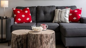 A modern living room with a grey sofa, red and white patterned pillows, a wooden coffee table with a bowl of popcorn and a mug, and a lamp on a side table. - Lovely 2 bedroom suite steps from The beach (Victoria)