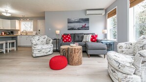 A modern living room with a grey sofa, patterned armchairs, and a red pouf. A wooden coffee table and a painting of a beach scene are also visible. - Lovely 2 bedroom suite steps from The beach (Victoria)