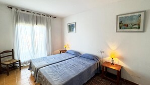 A room with two beds, a chair, a small table, and a painting on the wall.