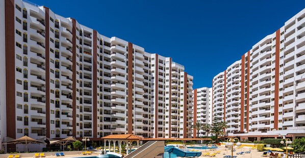 A high-rise hotel with multiple balconies, a swimming pool area with sun loungers, and a covered pavilion.