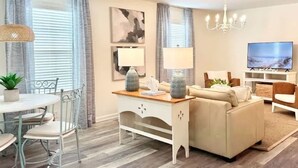 A modern living room with a sofa, dining table, and chairs. There is a TV and a chandelier. - Sandy Shores - Near Redfern (St. Simons Island)