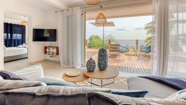 A modern hotel room with a bed, a TV, a balcony with outdoor furniture, and a view of the sea.