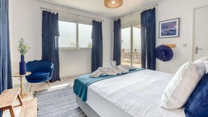 A modern hotel room with a large bed, a blue chair, a small table, and a view of the outdoors.