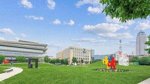 Une zone verdoyante avec un bâtiment moderne, une sculpture et, au loin, une haute tour.