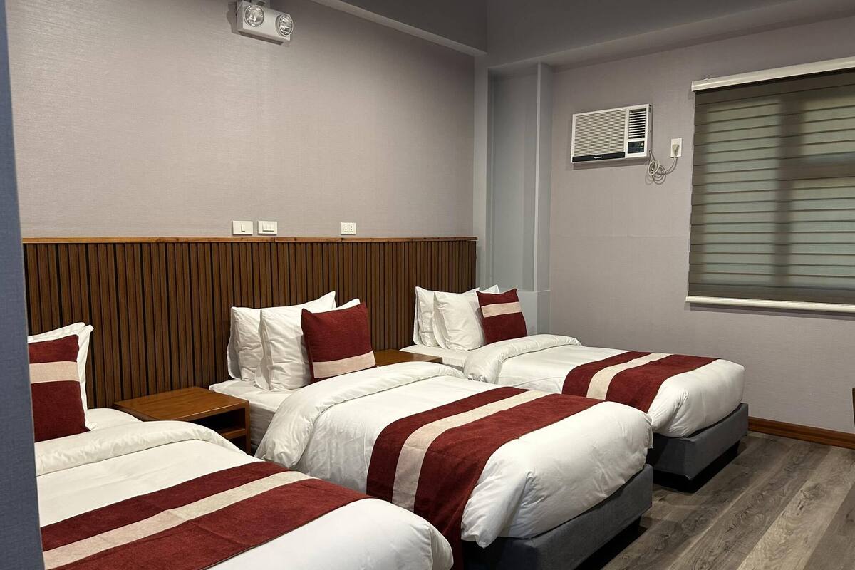 Hotellihuone, jossa on kaksi sänkyä, puinen pääty, ikkuna, jossa on sälekaihtimet, ja seinälle asennettu ilmastointilaite.