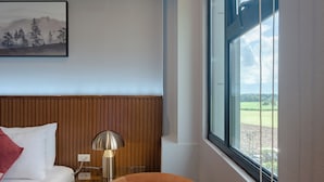 A hotel room with a wooden headboard, a bed with white bedding, a round orange chair, a bedside table with a lamp, and a window with a view of a green landscape. - Kekehyu Business Hotel (Valencia)