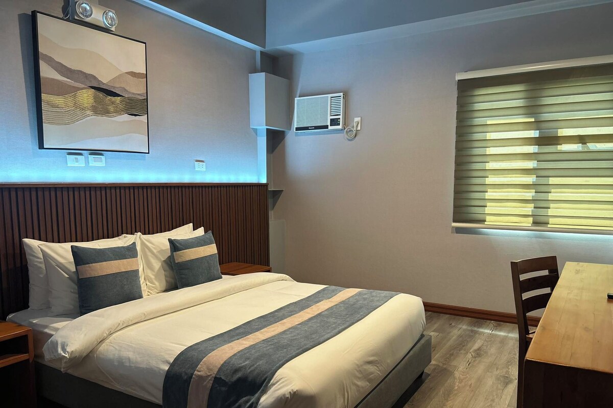 Quarto bem arrumado com cama, mesas de cabeceira, escrivaninha e janela com persianas.