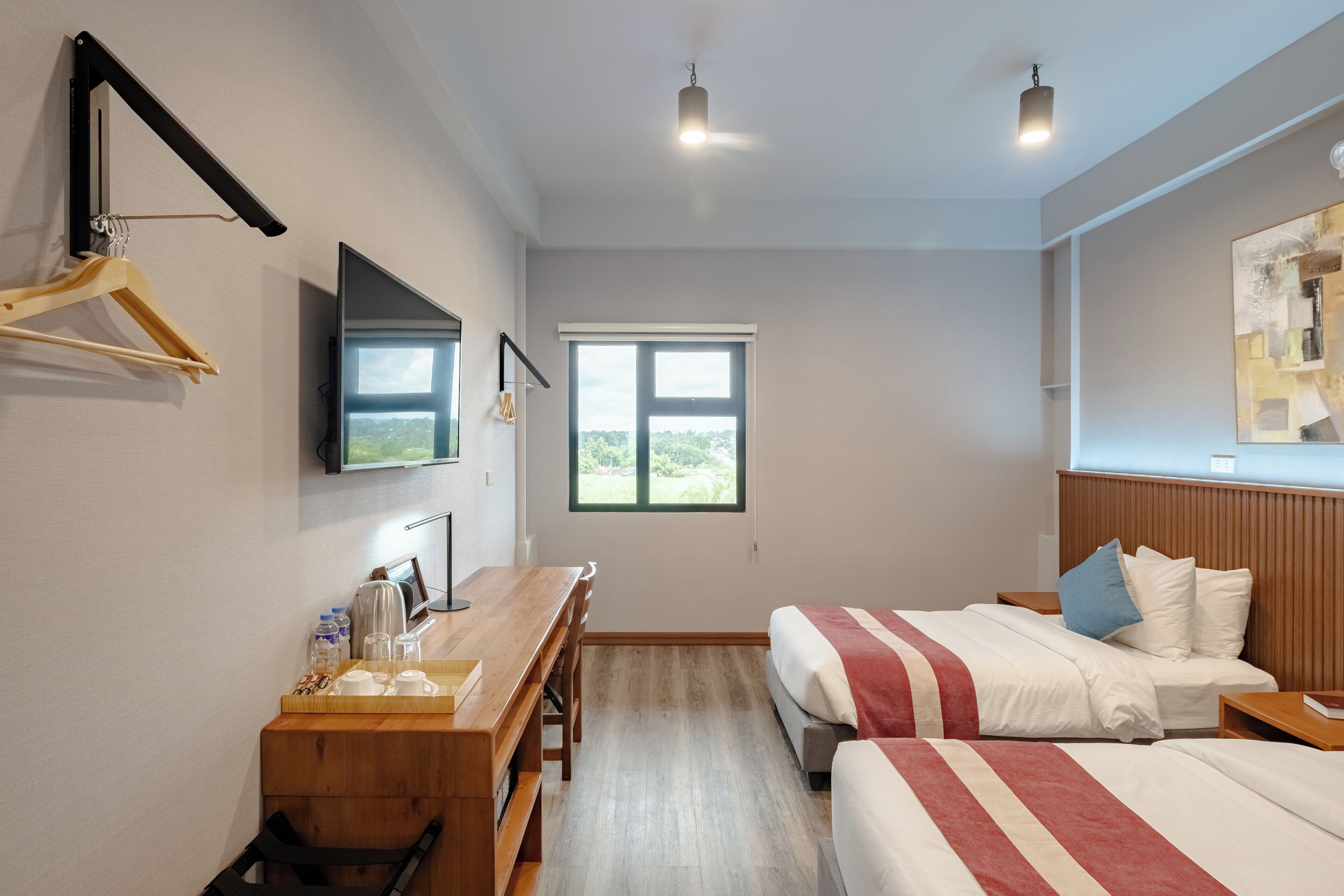 Habitación de hotel con un escritorio de madera, una cama con manta a rayas, un televisor montado en la pared y una ventana con vistas a zonas verdes.