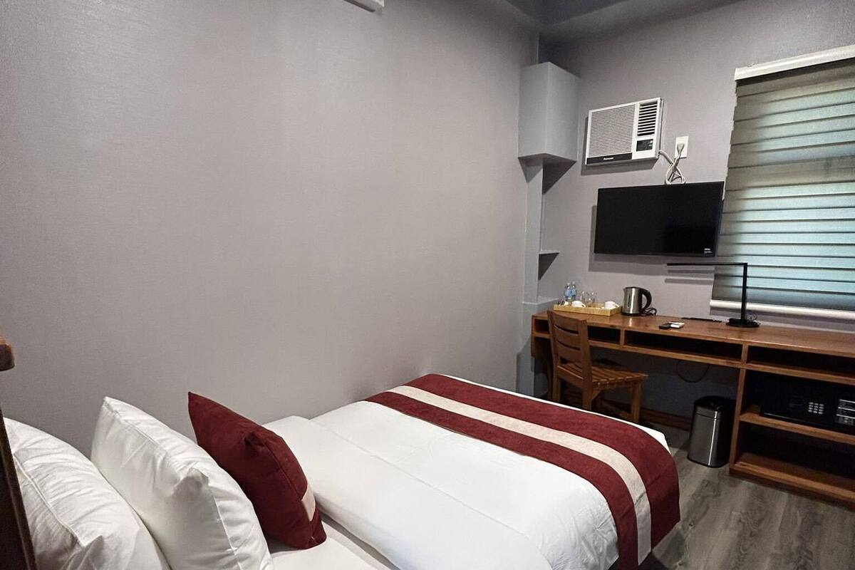 Hotellihuone, jossa on sänky, työpöytä, televisio ja ikkuna, jossa on sälekaihtimet.