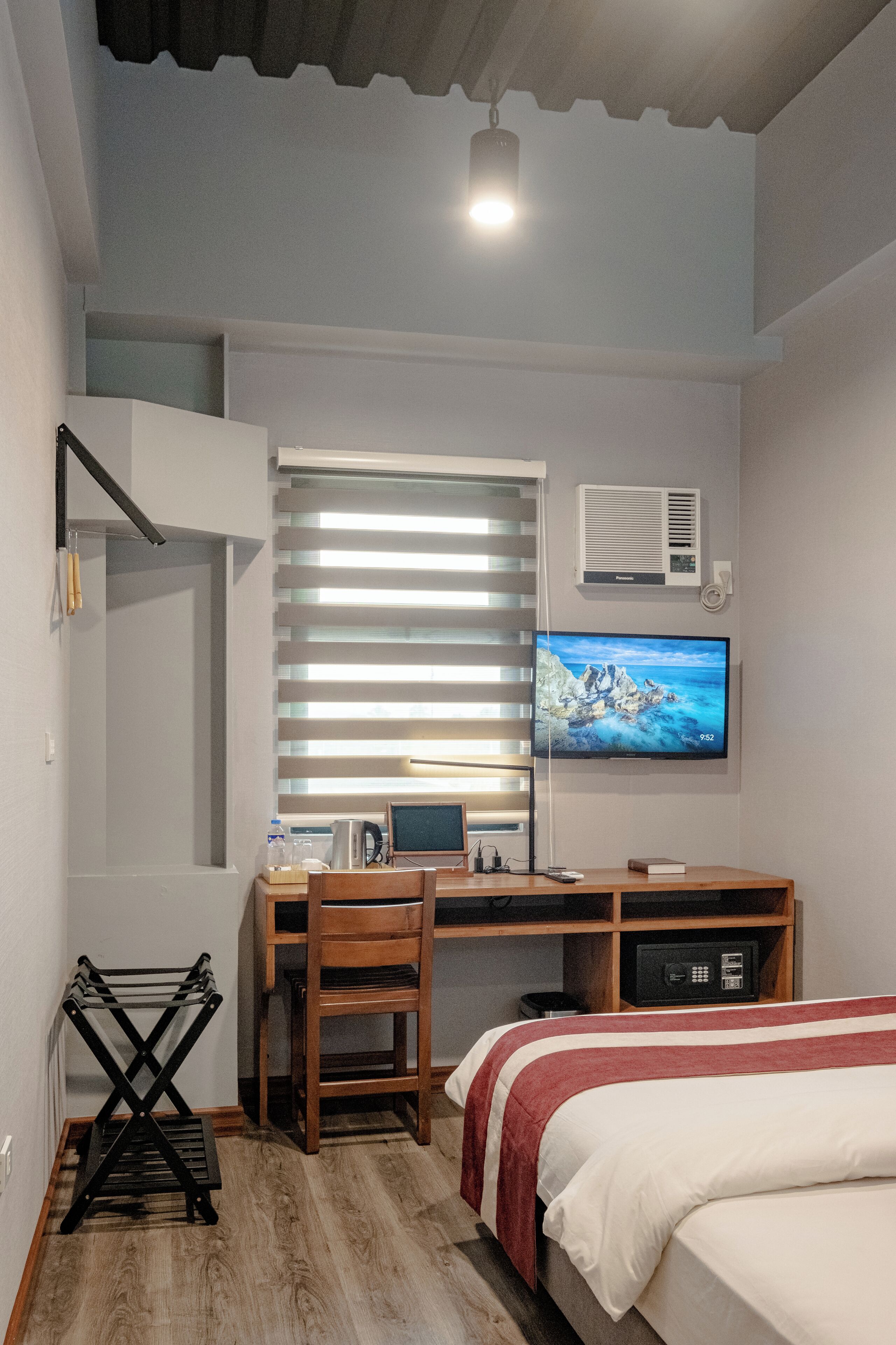 Una camera da letto con un letto, una scrivania con un computer, una sedia, una televisione e una finestra con persiane.