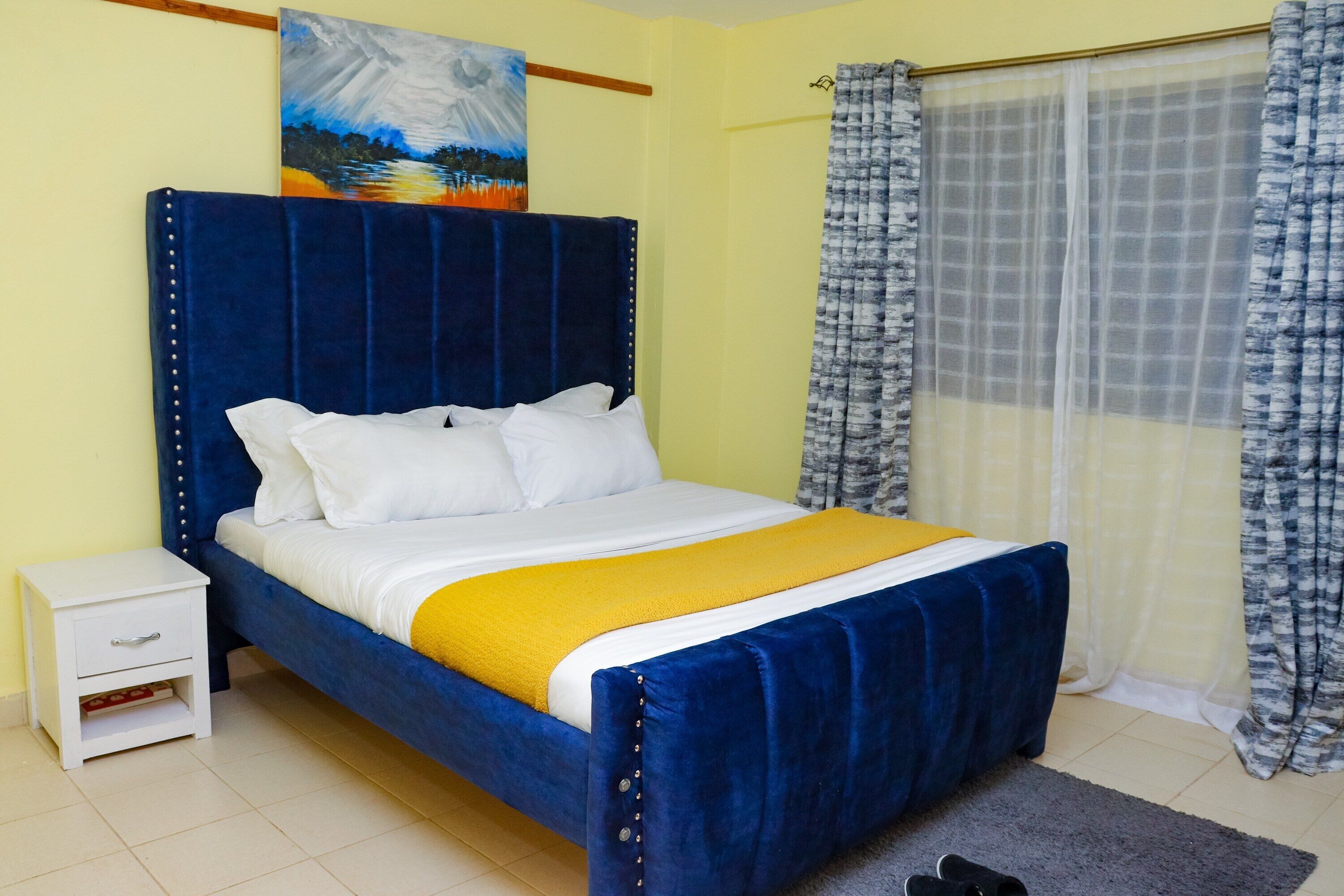 Un dormitorio con una cama azul, almohadas blancas, una colcha amarilla, una mesita de noche blanca y un cuadro en la pared.
