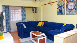 Uma sala de estar com um sofá azul, uma mesa pequena branca e uma televisão. Há duas pinturas na parede.
