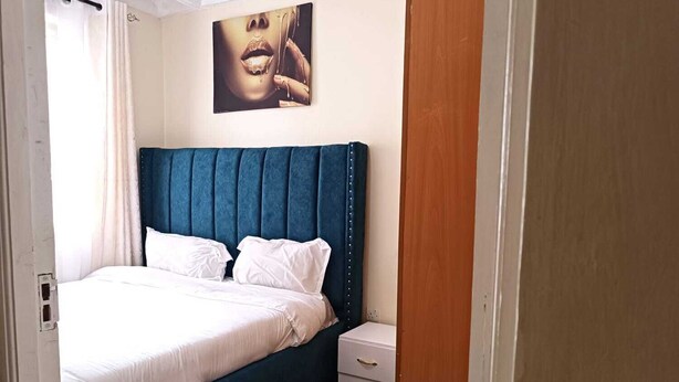 Quarto com cabeceira estofada em azul, roupa de cama branca e uma pequena mesa de cabeceira com gavetas.