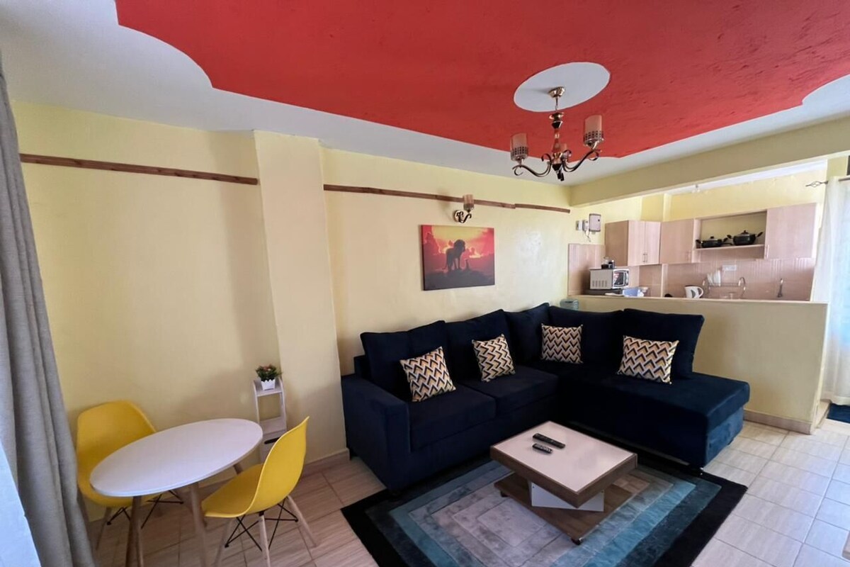 A living room with a dark blue sofa, yellow chairs, a small white table, and a kitchen in the background.