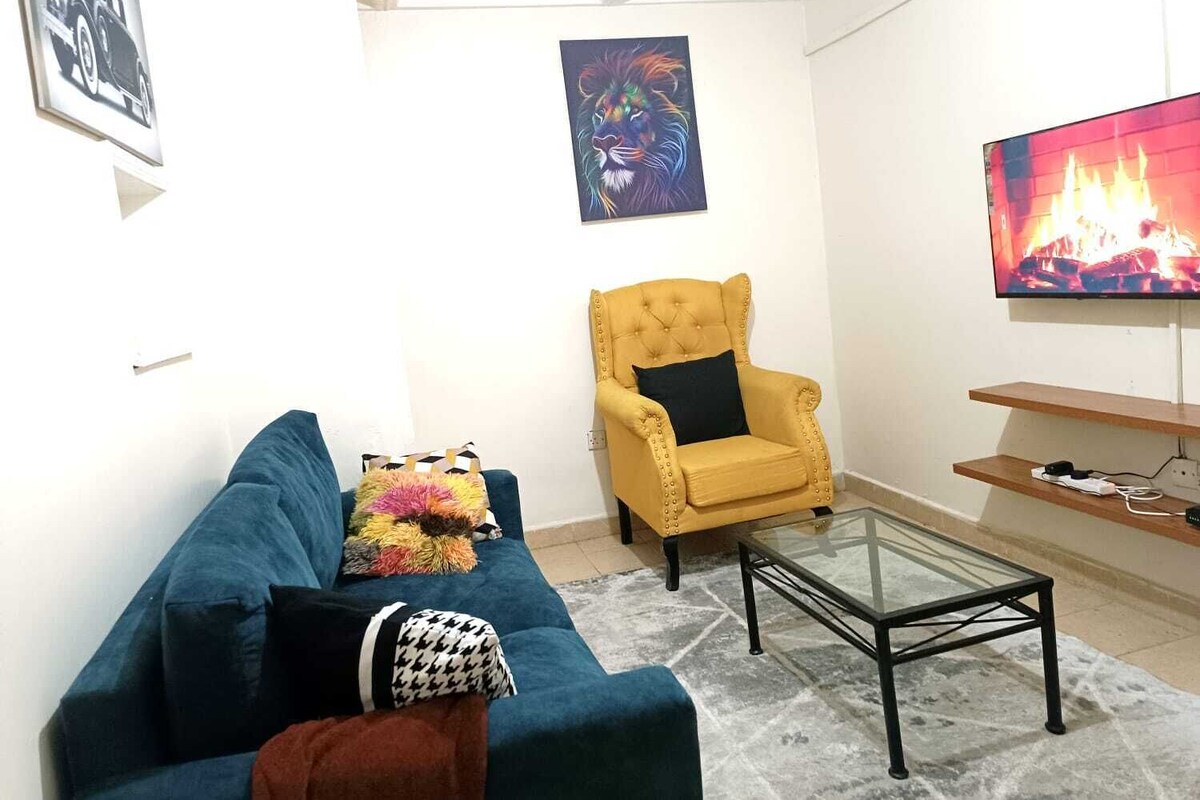 A living room with a blue sofa, a yellow armchair, a glass coffee table, and a TV displaying a fire.