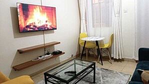Uma sala de estar com uma TV exibindo uma lareira, uma mesa de centro de vidro, cadeiras amarelas e um sofá.