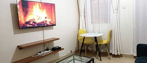 A living room with a TV displaying a fire, a glass coffee table, yellow chairs, and a sofa.