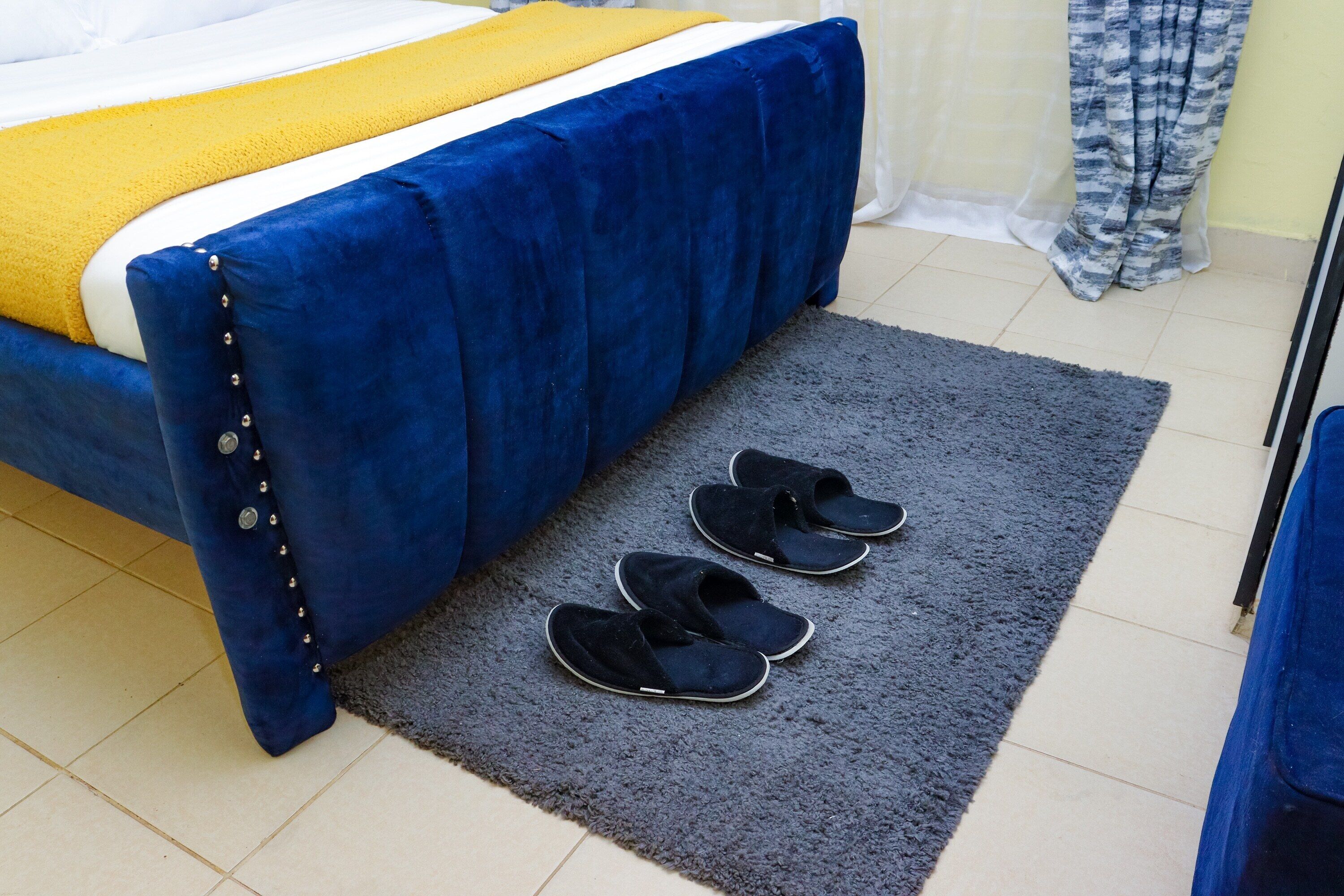 Een bed met een blauwe hoes en een gele deken, een grijs vloerkleed en twee zwarte pantoffels.