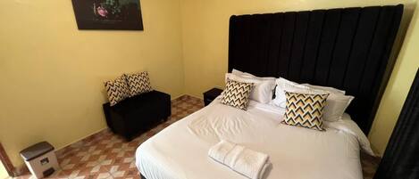 A hotel room with a large bed, a black headboard, and a small sofa. There is a trash bin and a painting on the wall.
