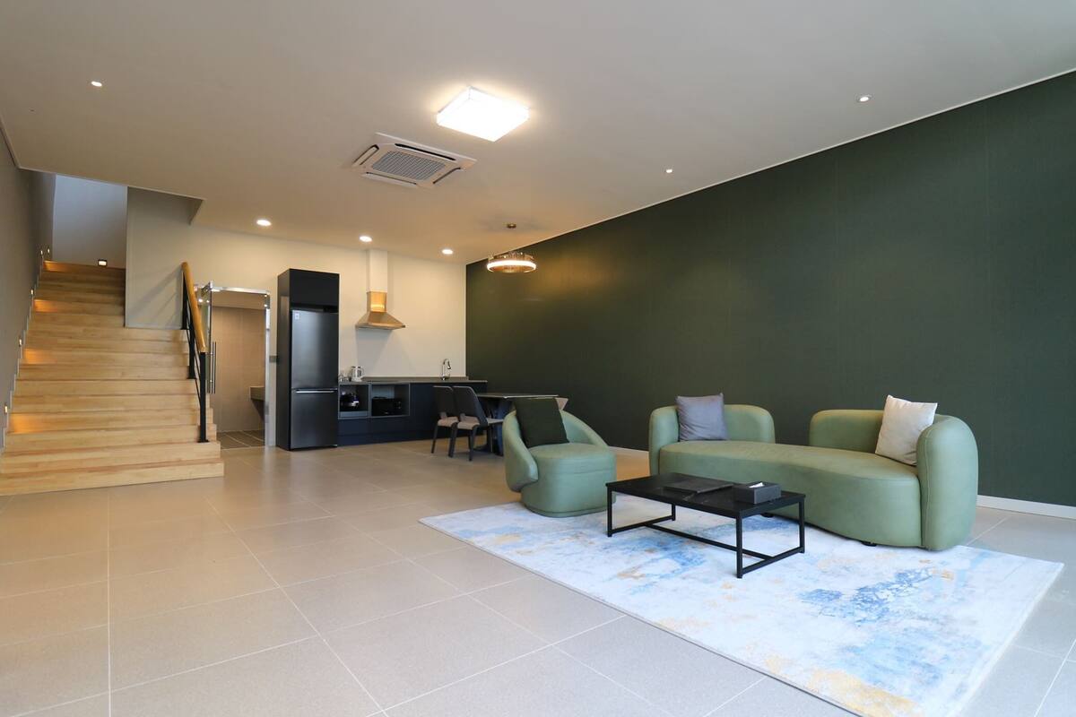 Una sala moderna con un sofá, sillones y una mesa de centro. Se ve un área de cocina al fondo, con un refrigerador y una estufa.