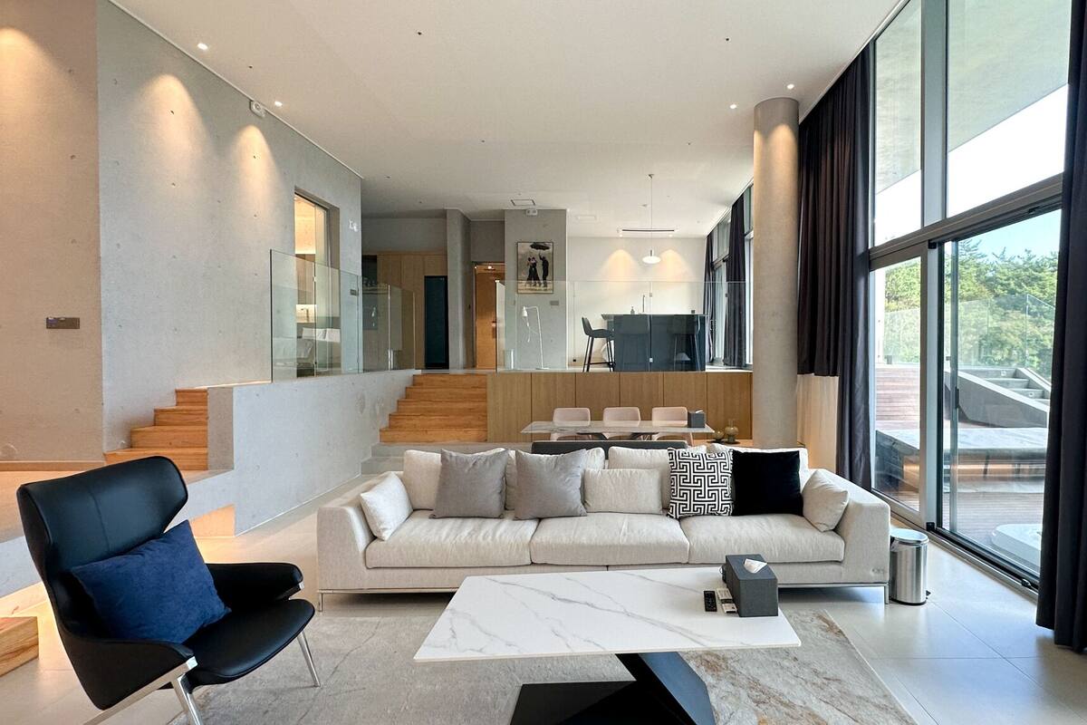Una sala de estar moderna con un sofá, una mesa de centro y una silla. Se ve una escalera que conduce a un nivel superior con un comedor.