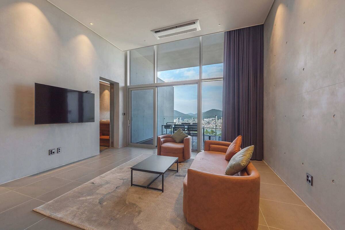 Una sala de estar moderna con un televisor de pantalla plana, un sofá, una mesa de centro y vistas a un paisaje urbano a través de grandes ventanales.