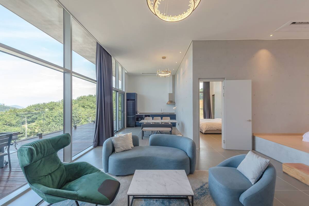 Una sala de estar moderna con ventanas amplias, un sillón verde, dos sofás azules y una mesa de centro de mármol.