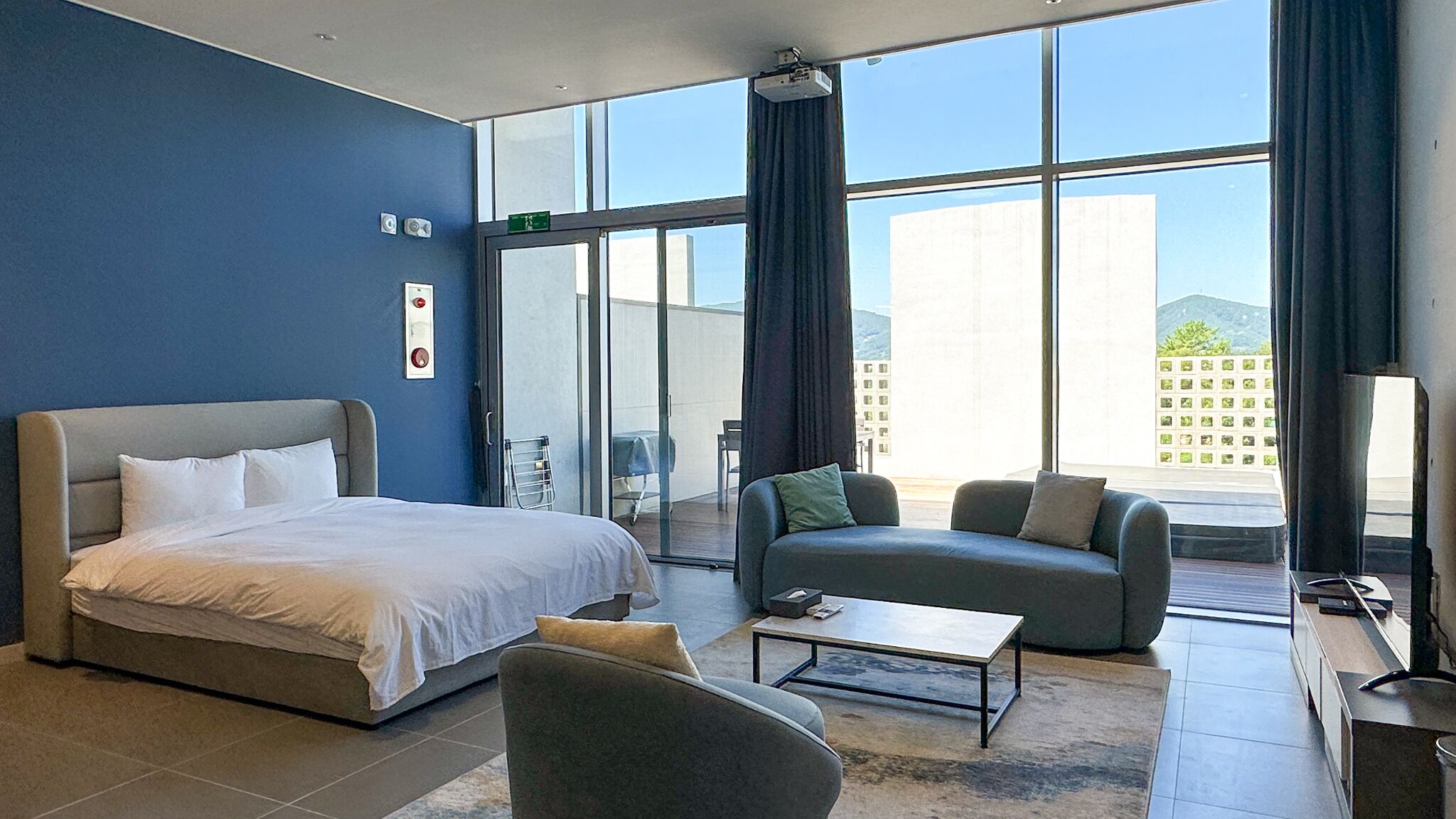 Quarto de hotel moderno com cama, sofá, mesa de centro e vista para a cidade através de amplas janelas.