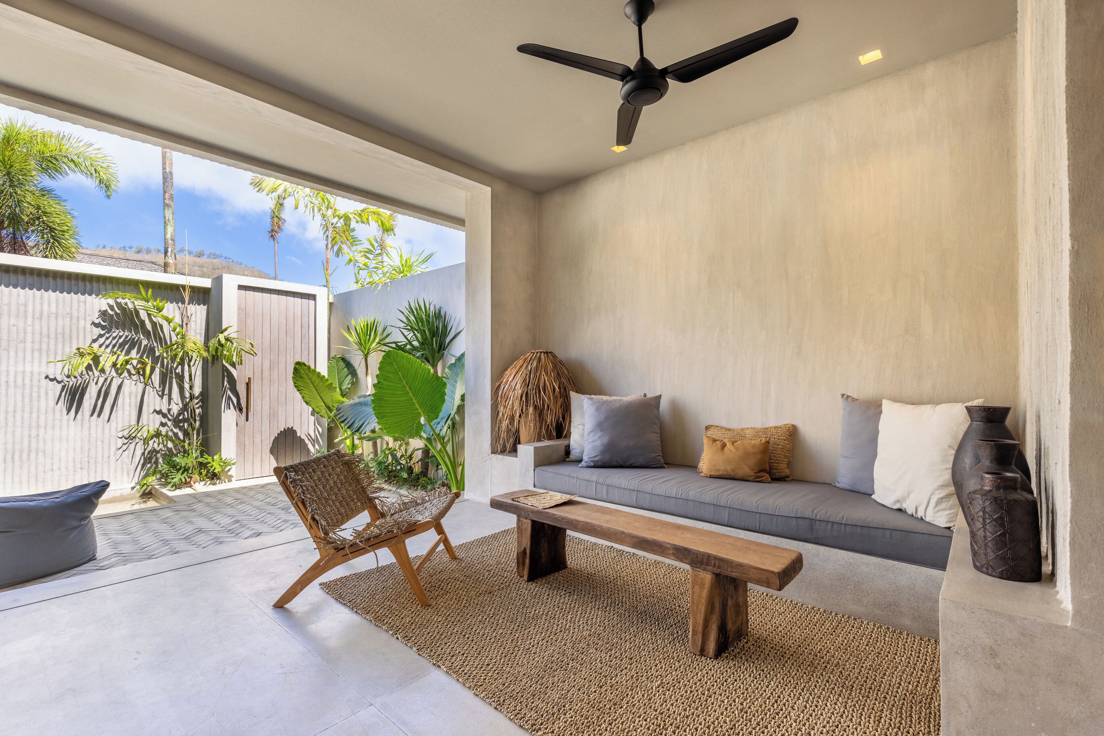 A modern living room with a sofa, bench, and chair, a ceiling fan, and large windows overlooking a garden.