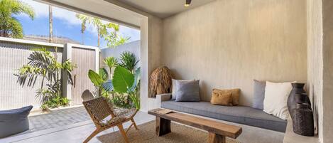 A modern living room with a sofa, bench, and chair, a ceiling fan, and large windows overlooking a garden.