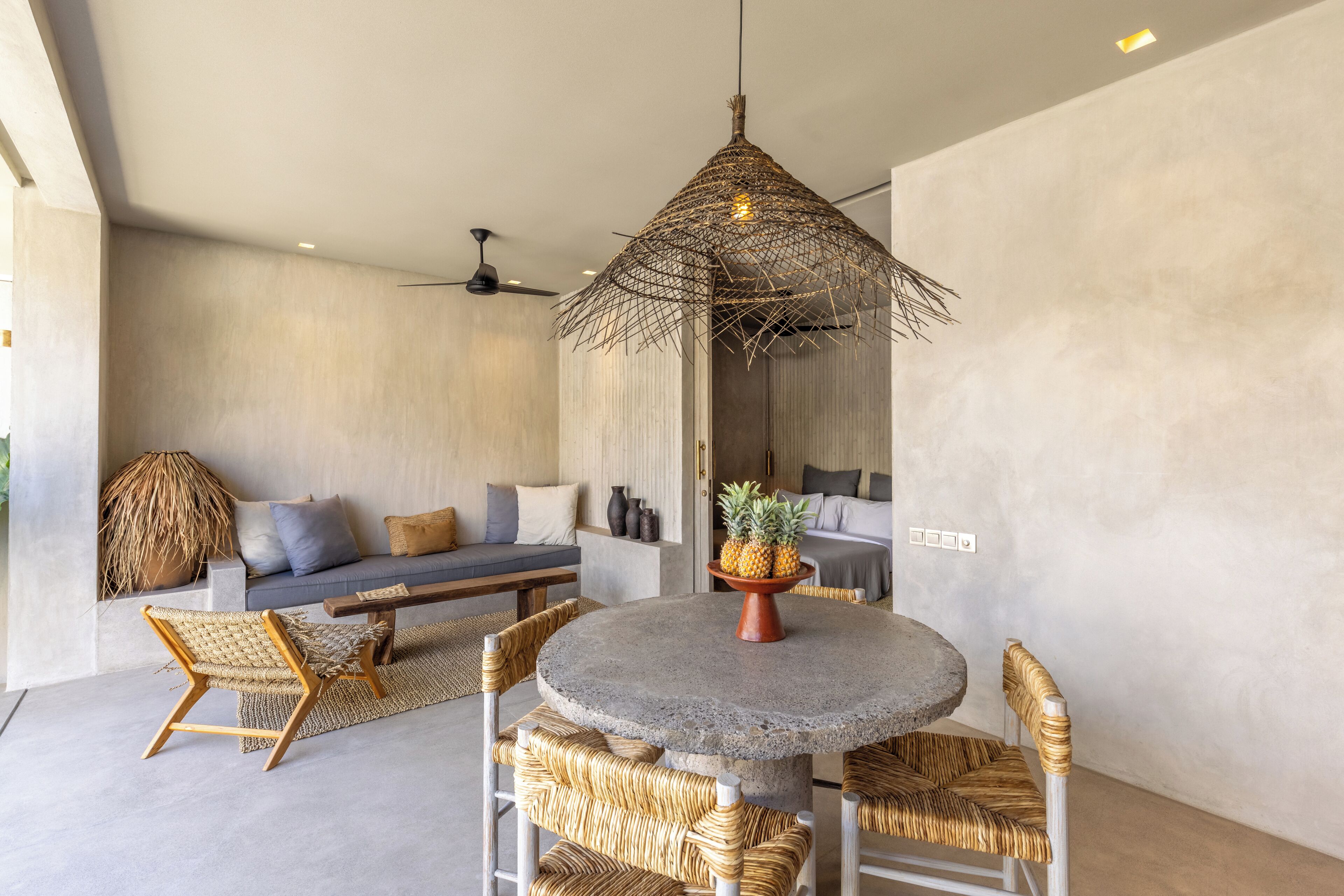 A modern interior with a round stone table, wicker chairs, a woven ceiling light, and a sofa with cushions.