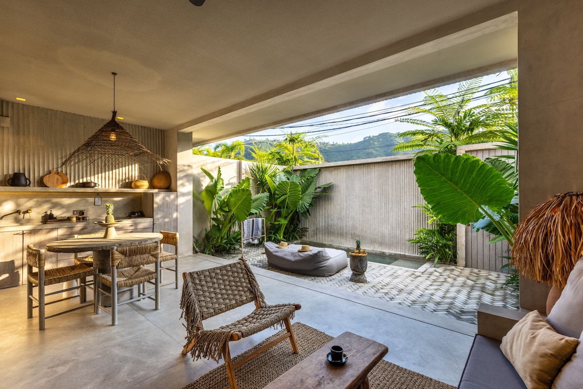 A modern kitchen with a dining area, a woven chair, and a view of lush greenery.