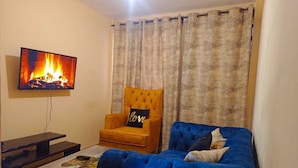 A living room with a television displaying a fireplace, a blue sofa, an orange armchair, and a coffee table. - Lux Suites Trm drive Apartments Roysambu (Nairobi)