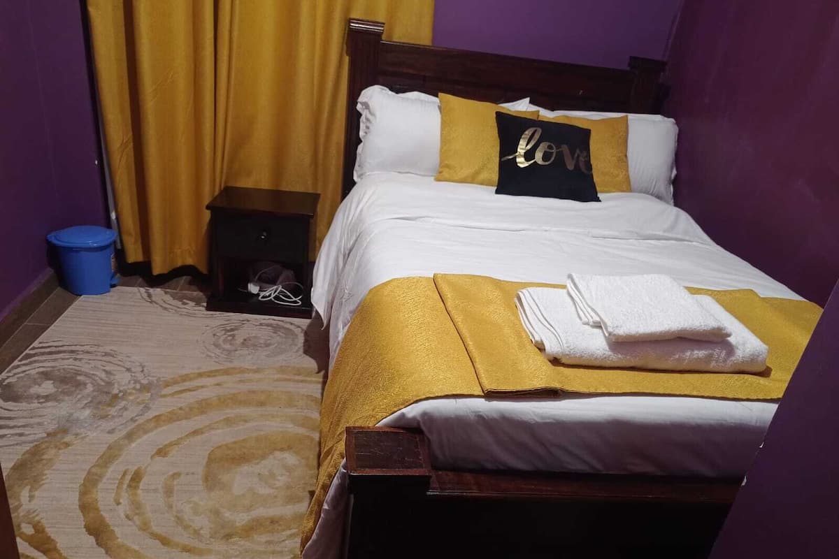 Un lit bien fait, avec une literie blanche et dorée, un oreiller orné du mot « amour », une poubelle bleue et un tapis à motifs.