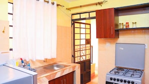 A compact kitchen with a gas stove, a sink, and a window with curtains. - Lux Suites Trm drive Apartments Roysambu (Nairobi)