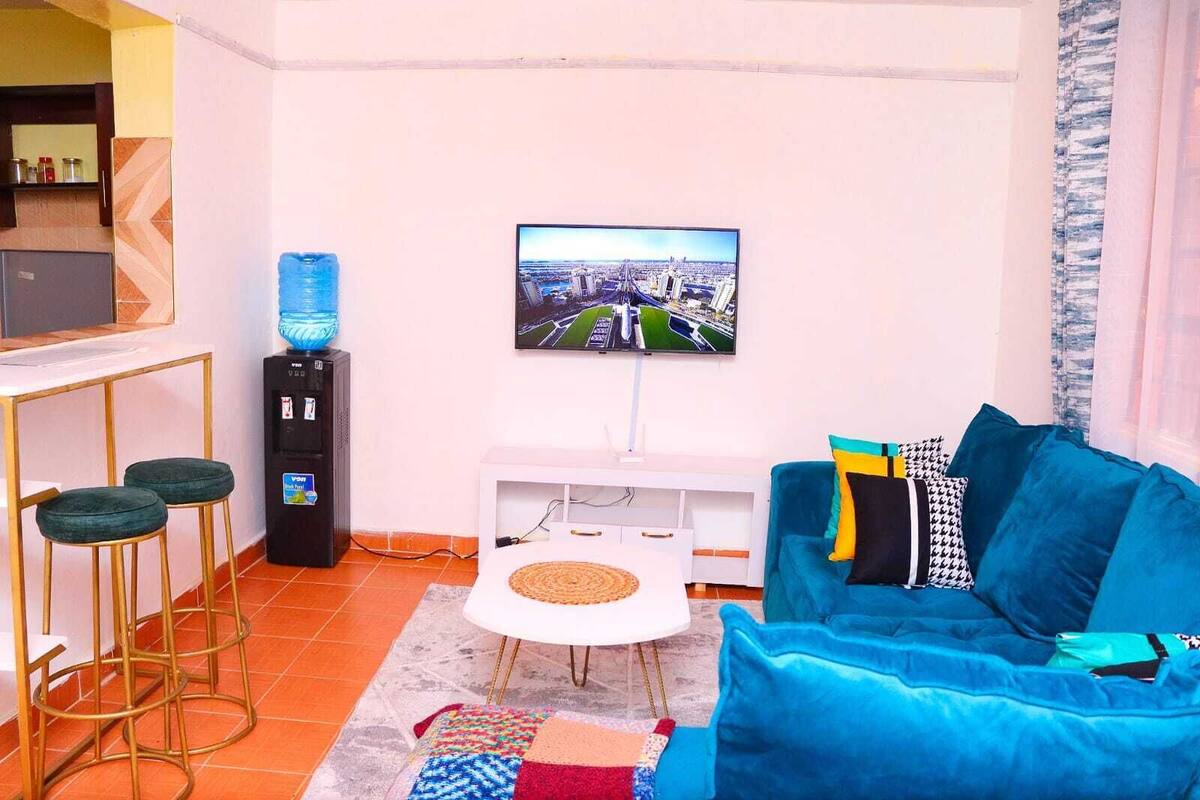 Un salon avec un téléviseur fixé au mur, un canapé bleu, une petite table blanche et un distributeur d’eau.