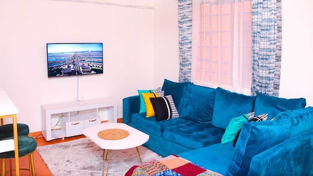 A living room with a blue sofa, a white TV stand, and a colorful rug. - Lux Suites Trm drive Apartments Roysambu (Nairobi)