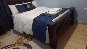 A bed with a wooden frame, white bedding, and a blue blanket. There is a pillow with the word "love" on it. - Lux Suites Trm drive Apartments Roysambu (Nairobi)