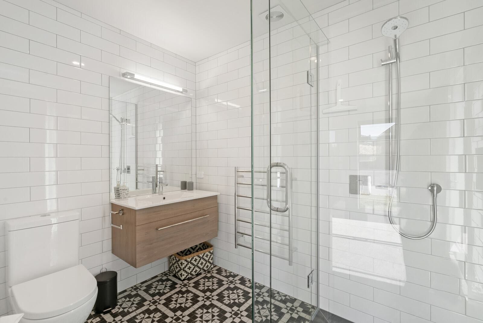A modern bathroom with white subway tiles, a glass shower enclosure, a wooden vanity, and patterned floor tiles.