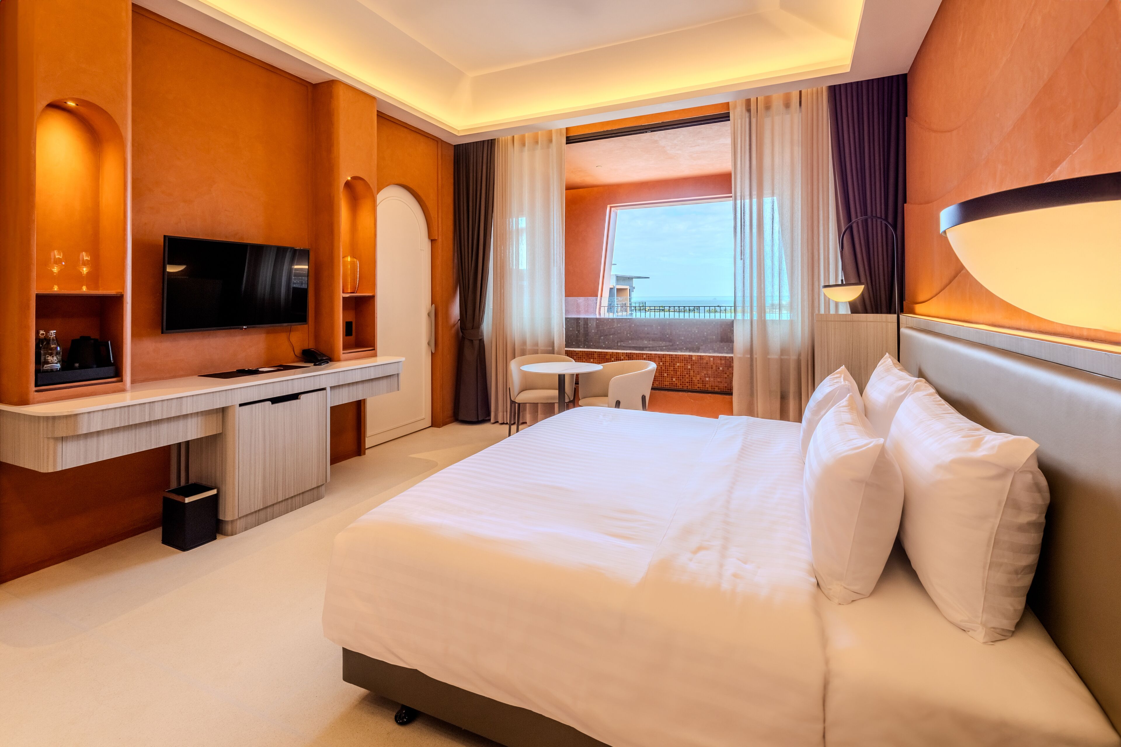 Habitación de hotel con una cama grande, un escritorio con televisor de pantalla plana, una mesita pequeña y vistas al mar a través de amplios ventanales.
