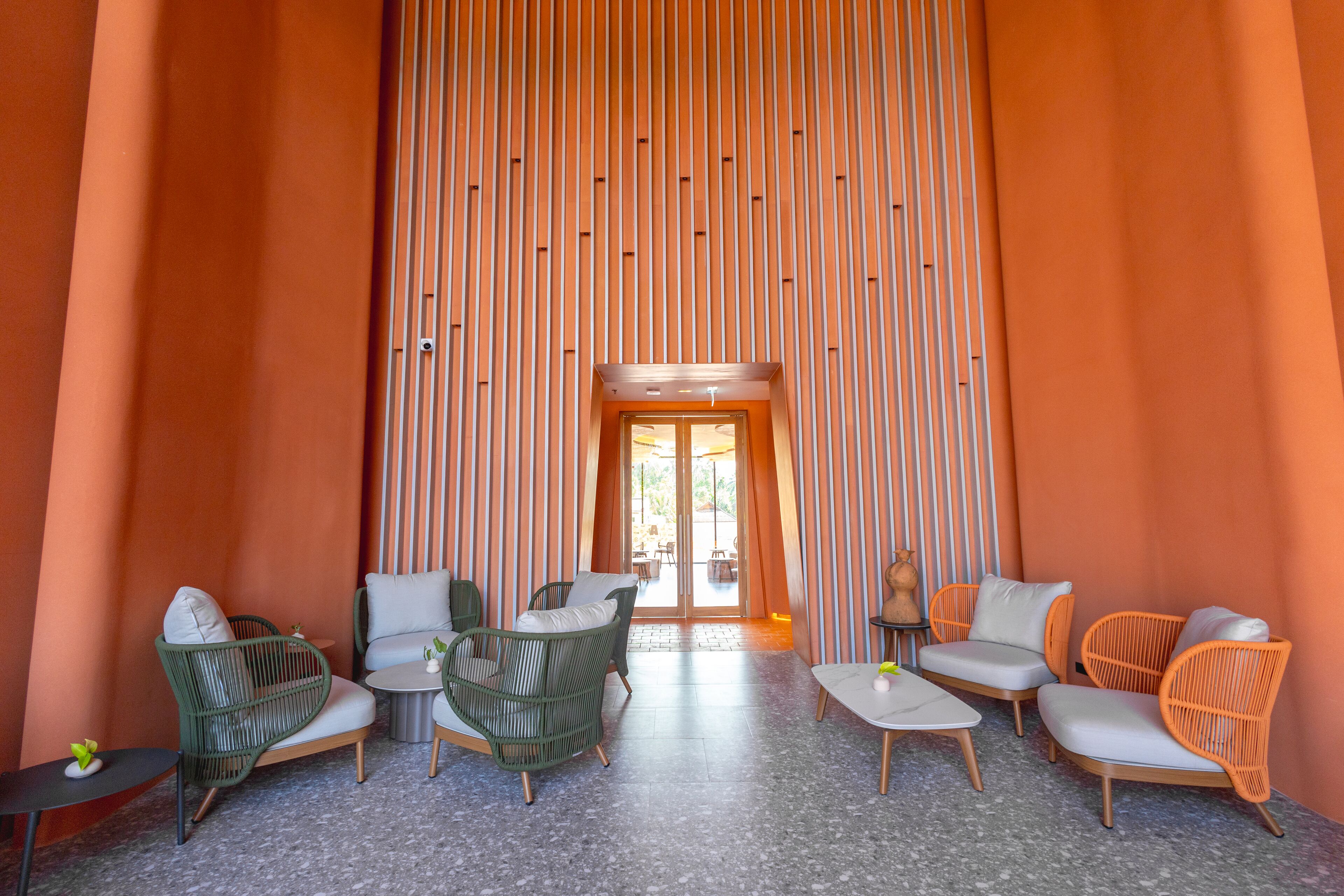 Un espace intérieur spacieux doté d’un haut plafond, aménagé avec des fauteuils en osier vert et orange, une petite table ronde et une porte donnant sur un espace extérieur.