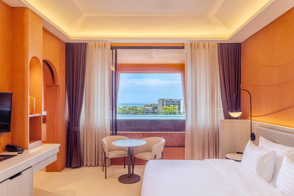 Habitación de hotel con cama, escritorio, silla, mesita, televisor y ventana grande con vistas a la ciudad.