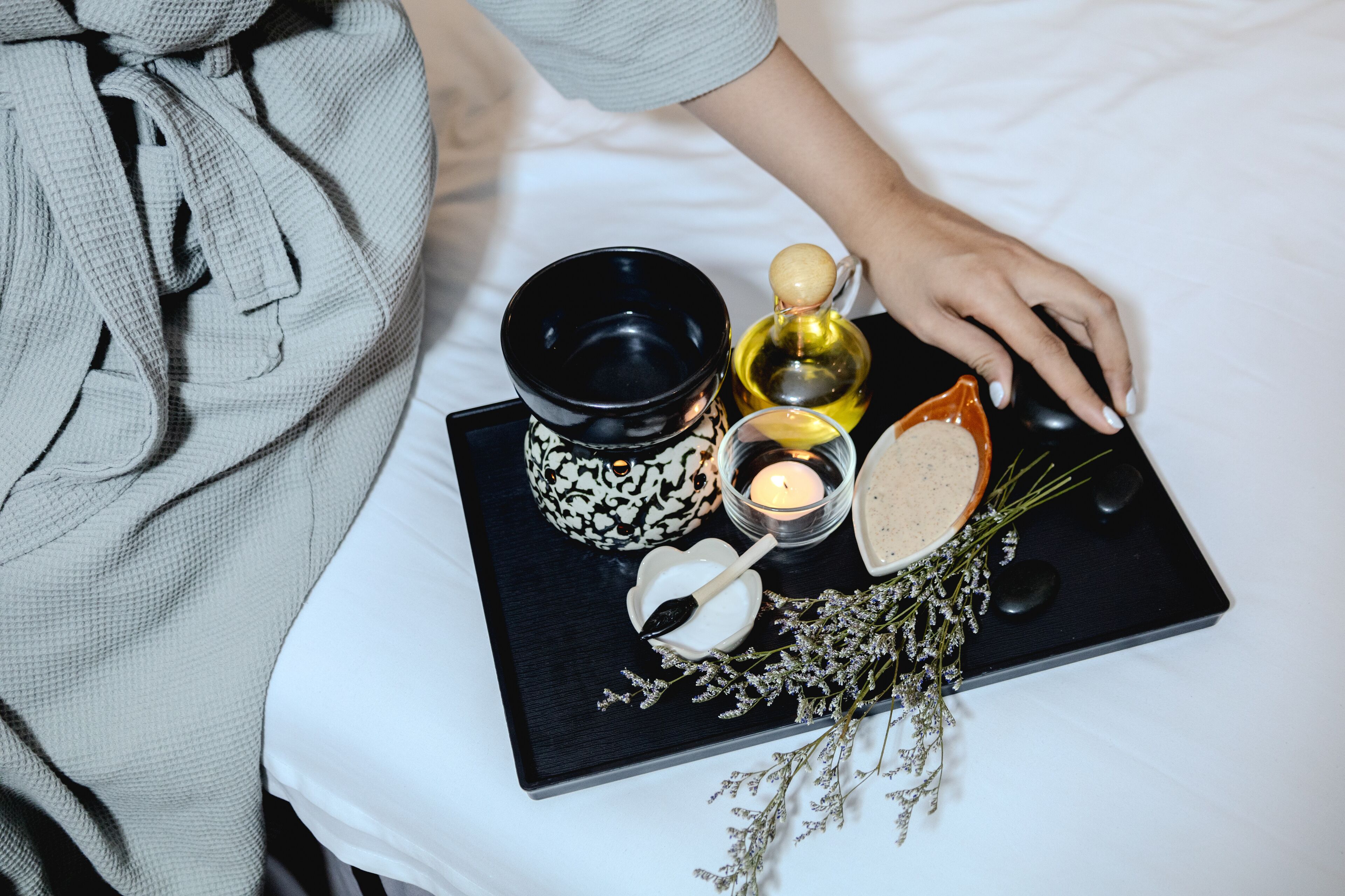 A tray with a black bowl, a candle, a bottle of oil, a bowl of salt, and a sprig of lavender on a white bed.
