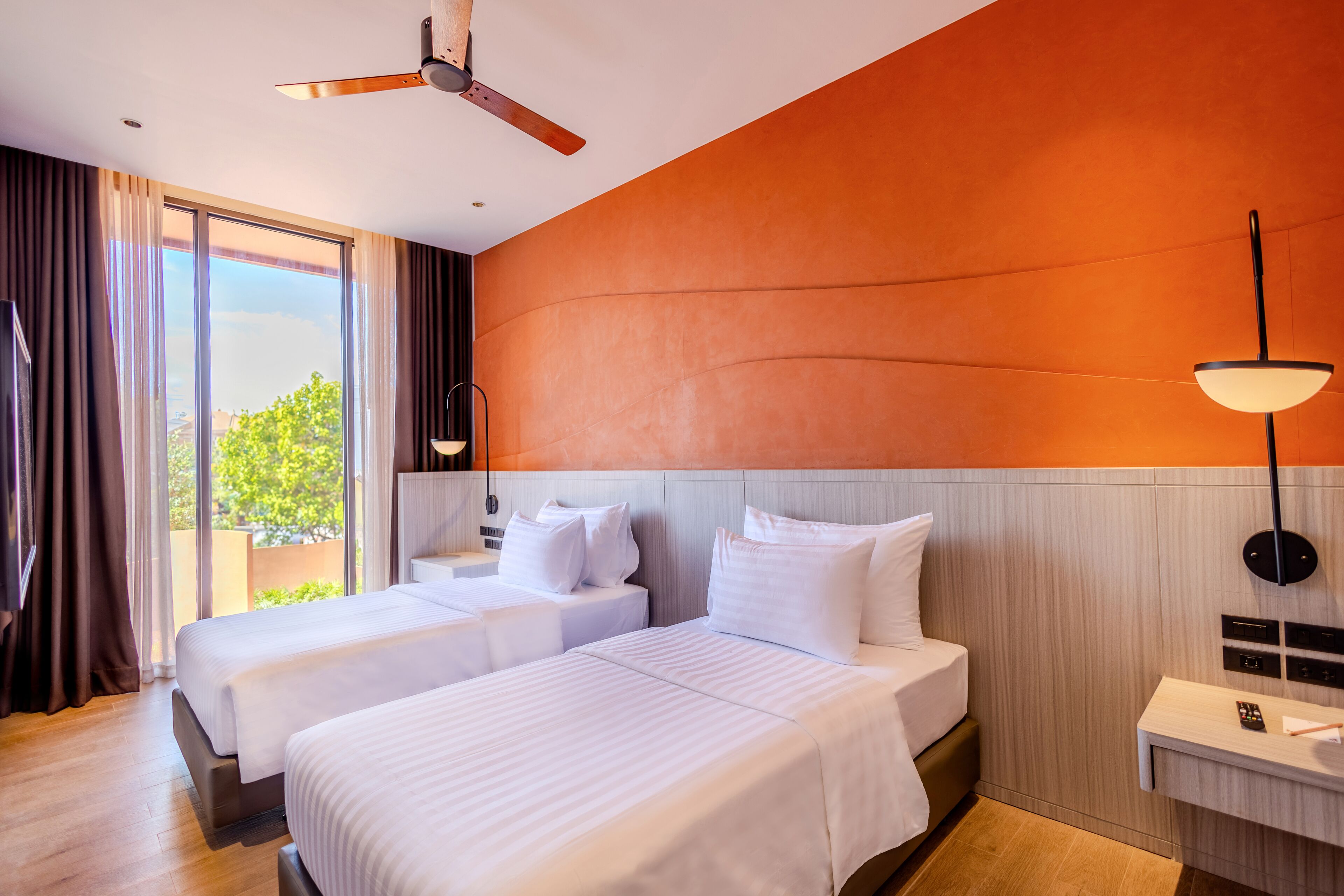 Une chambre d’hôtel avec deux lits, un ventilateur de plafond, une grande fenêtre avec des rideaux, une table de chevet et une lampe fixée au mur.