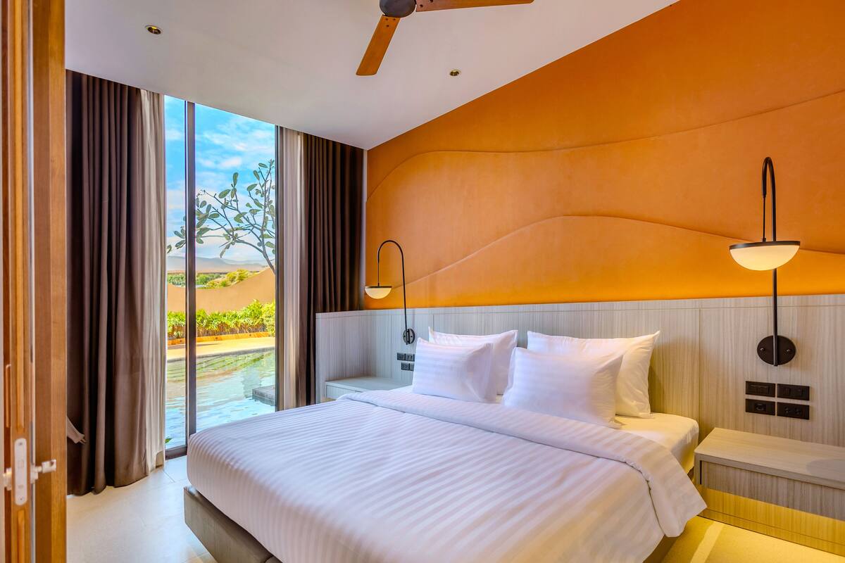Habitación de hotel con una cama grande, una mesita de noche, una lámpara de pared y vistas a una piscina y al paisaje.