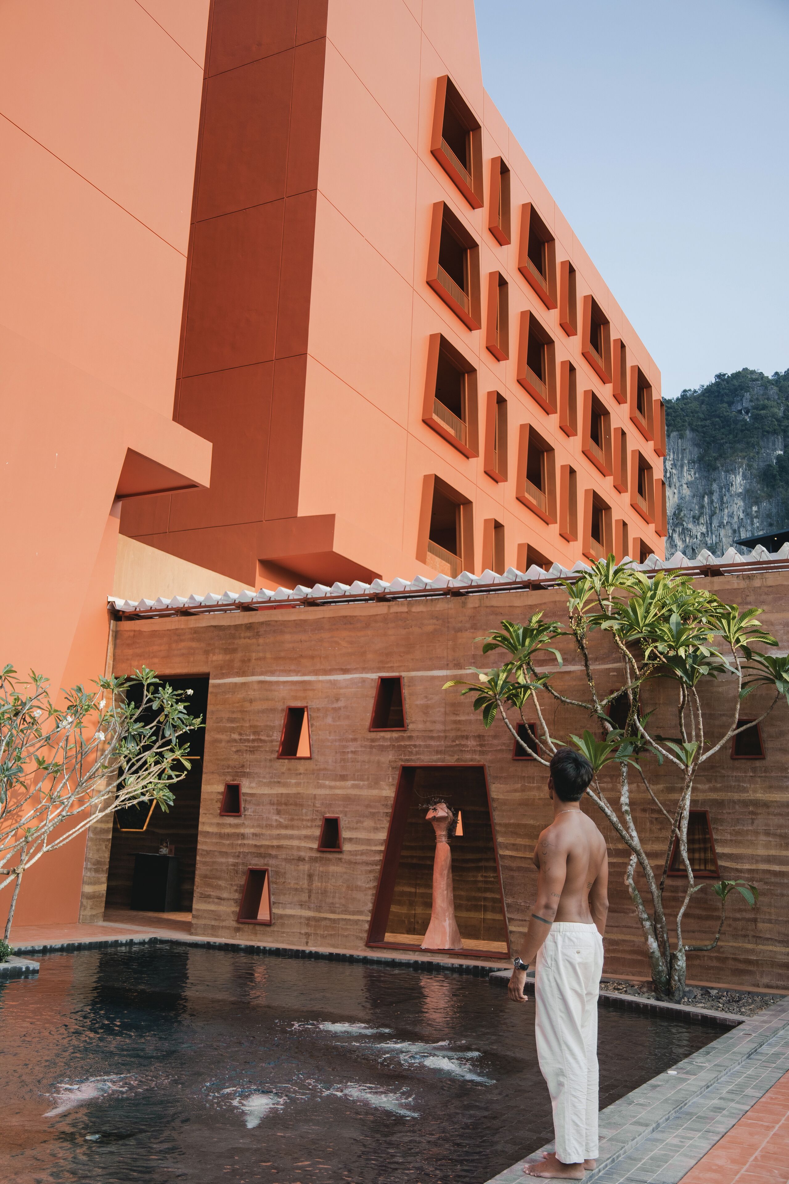 A man standing by a pool with a building featuring geometric patterns and a statue in the background.