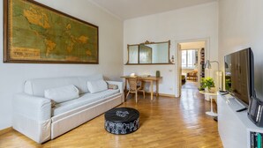 Un salon avec un canapé blanc, un pouf rond, une grande carte du monde, un miroir, un bureau et un téléviseur.